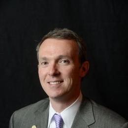 Man with dark grey suit with white dress shirt and purple tie in front of black background
